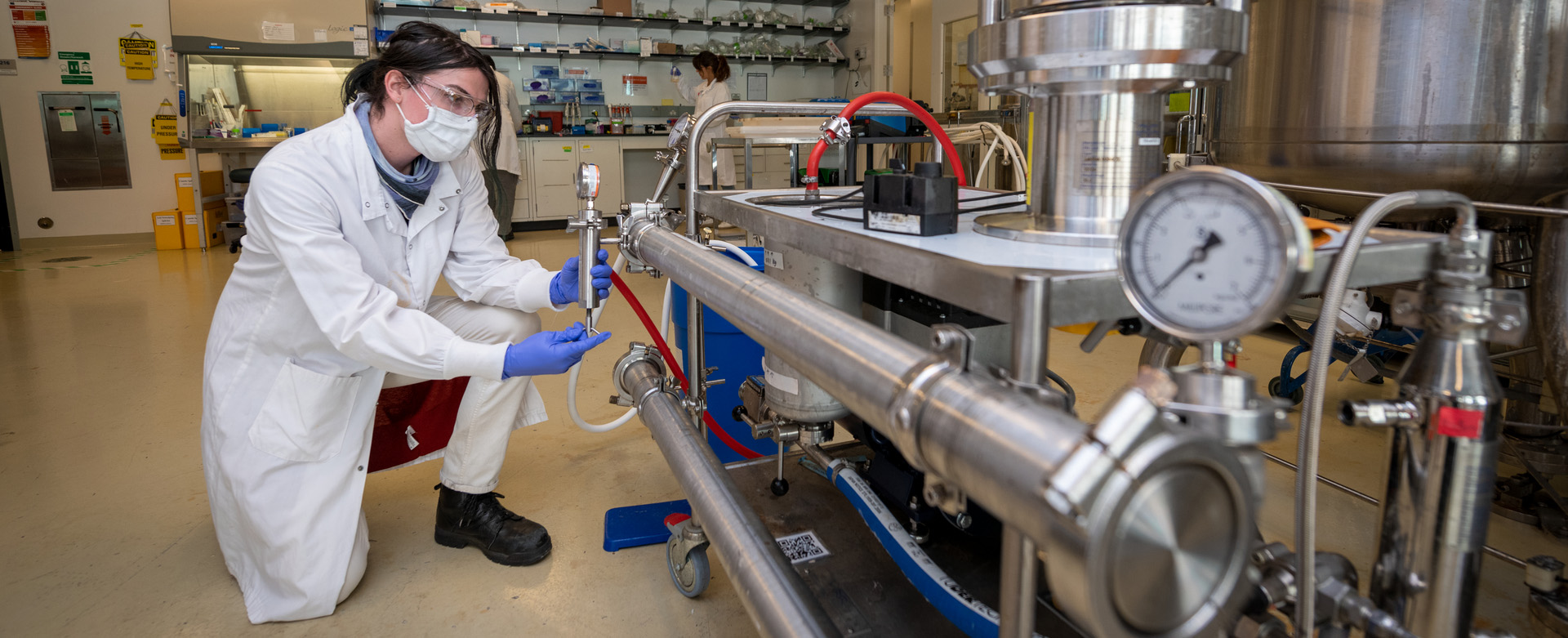 Dark-haired person in a lab coat kneels nextg to a piece of scientific machinery with values and gauges.