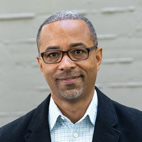Short haired person wearing glasses and a light collared shirt with a dark blazer.