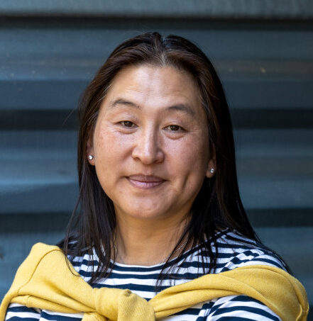 A person with long dark hair wearing a black and white striped shirt and a yellow cardigan tied around their shoulders poses.