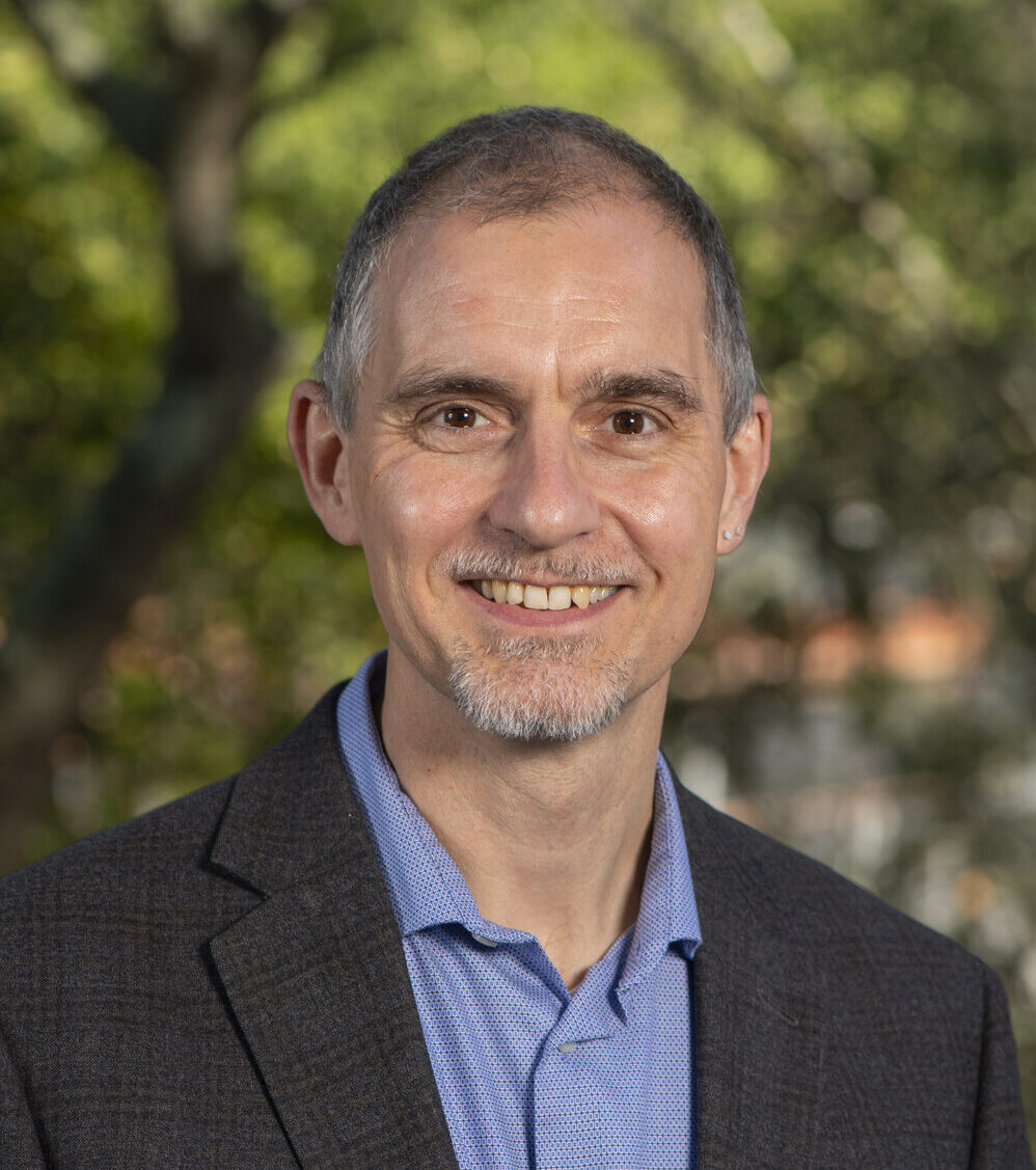 Person with short gray hair wearing a dark gray blazer over a light blue collared shirt.