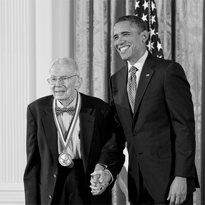 Two men in black suits looking forward. The person on the left is wearing a medal.