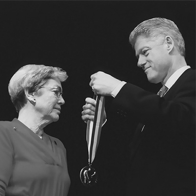 Two people in black suits. The person on the right is preparing to place a medal on the other person.