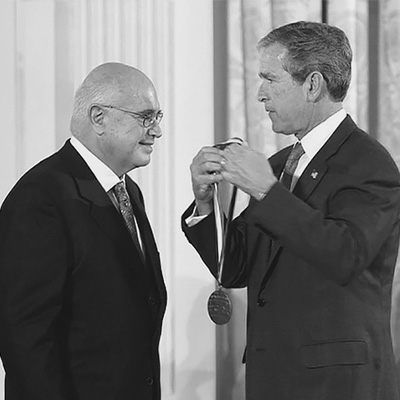 Two men in black suits. The person on the right is preparing to place a medal on the other person.