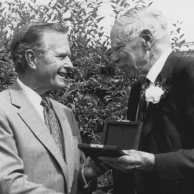Two people in dark suits. The person on the right is presenting a medal to the other person.