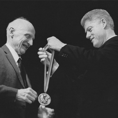 Two people in black suits. The person on the right is preparing to place a medal on the other person.
