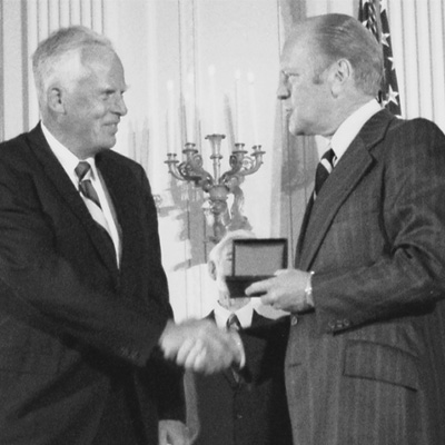 Two people in dark suits. The person on the right is presenting a medal to the other person.