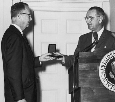 Two people in dark suits. The person on the right is presenting a medal to the other person.