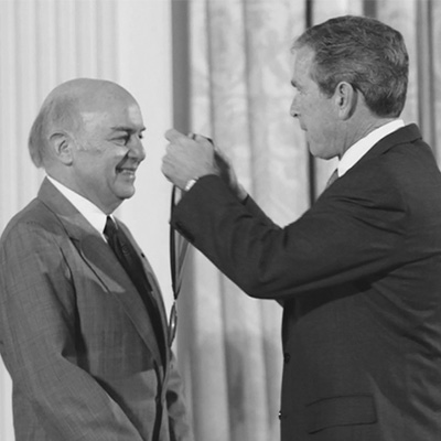 Two men in black suits. The person on the right is preparing to place a medal on the other person.