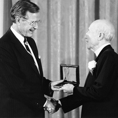 Two people in dark suits. The person on the left is presenting a medal to the other person.