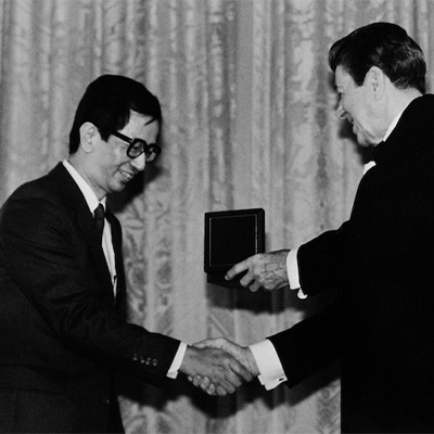 Two people in dark suits. The person on the right is presenting a medal to the other person.