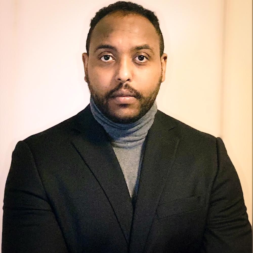 Mohammed Ombadi, a person with short black hair wearing a gray turtleneck and a black coat poses against a yellow backdrop.