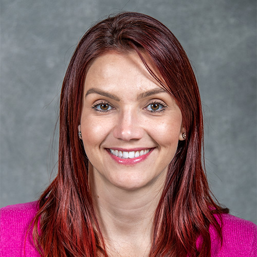 Person with long dark red hair wearing a pink shirt against a gray backdrop.