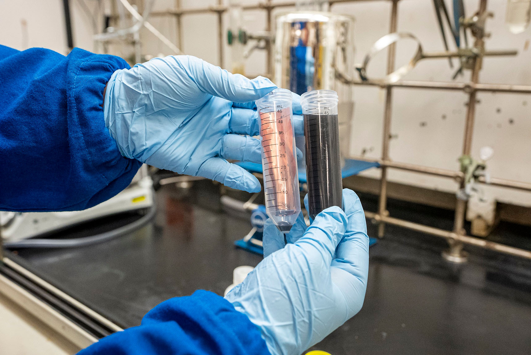 Two hands with blue gloves are golding tubes in a lab setting. One tube is filled with a piece of copper that has been cleaned of other components and the other with the alkaline solution used to dissolve the quick-release battery binder