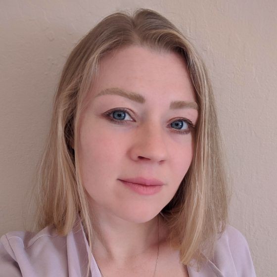 Person with medium-length blonde hair wearing a light purple blouse with a necklace.