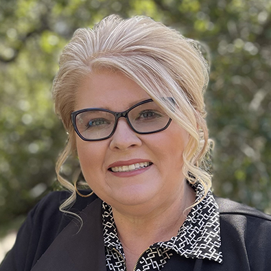 Blonde-haired person in glasses wearing a black blouse with floral patterns.