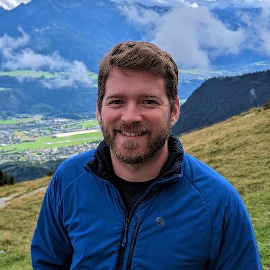 Person with short brown hair and a beard wearing a blue active jacket. He is standing on a grassy hillside, overlooking a town and mountains in the distance.