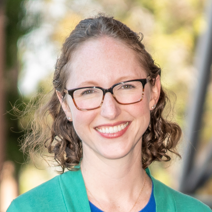 Corinne Scown, a person with shoulder-length brown hair wearing red and black glasses and a teal cardigan over a blue top.