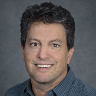 Person with short dark hair wearing a dark collared shirt against a gray photo drop background.