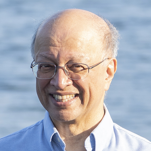 Smiling person with glasses wearing a blue collared shirt in front. The person is standing in front of water.