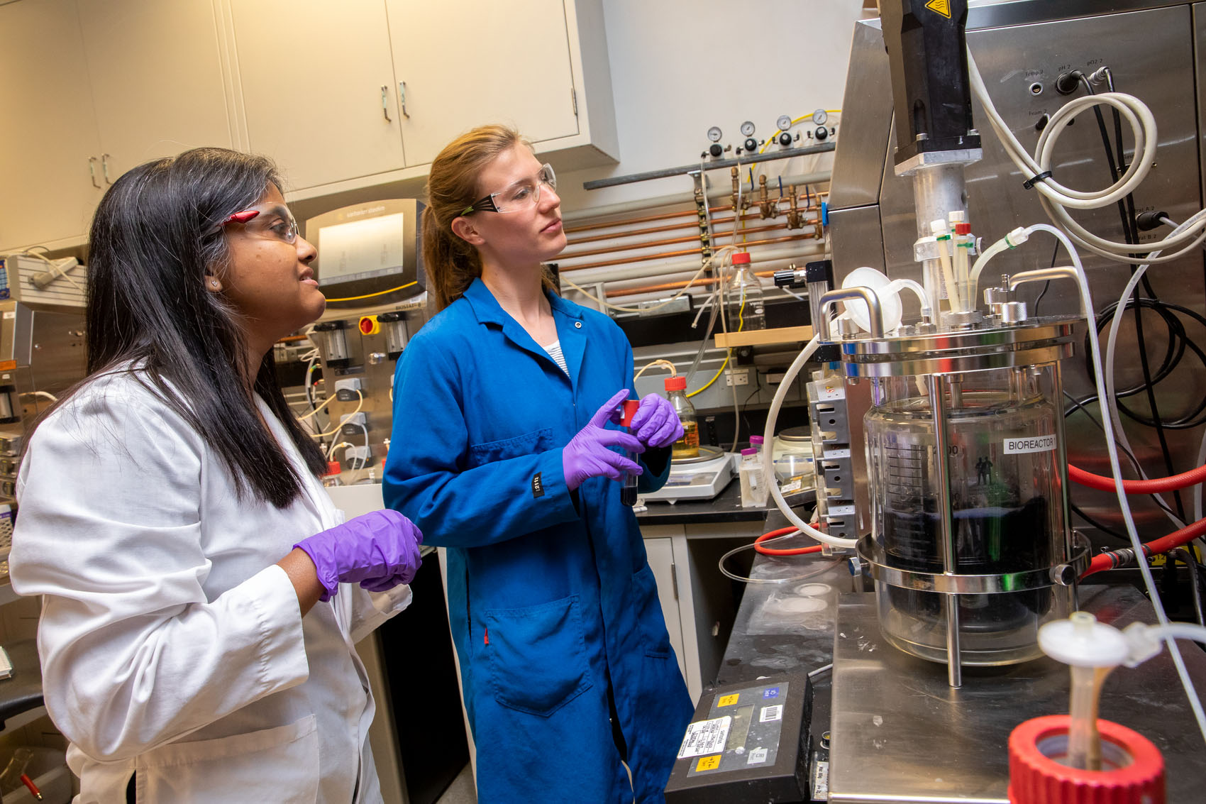 Two scientists wearing lab coats, gloves, and protective eye gear work in a lab.