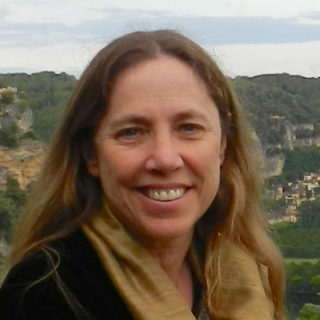 Person with long dark blonde hair wearing a black sweater and gold scarf. Photographed outdoors, with green vegetation and buildings in the background.