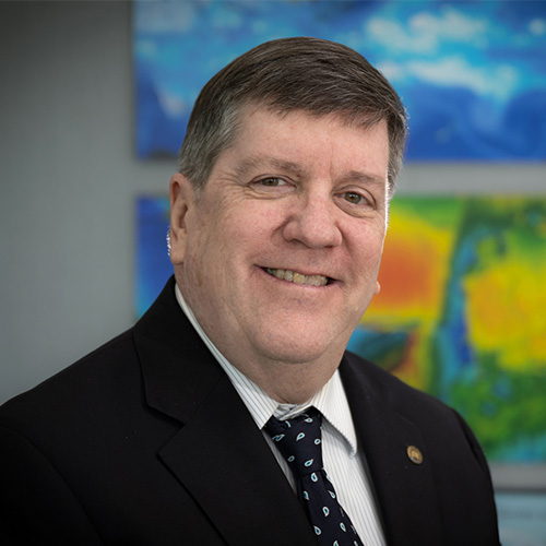 Smiling person with short gray hair wearing a black suit, navy tie, and a striped collared shirt photographed against climate models in the background.