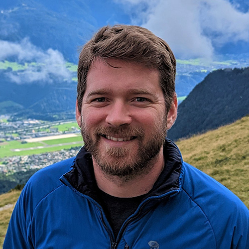 Person with short light brown hair wearing a blue jacket outdoors with green hills, trees, and mountains in the background. Below the hills is a town.