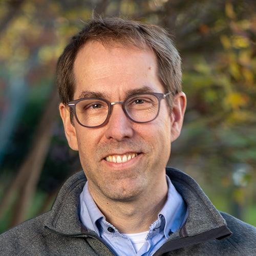 Person with short brown hair wearing glasses and a gray sweater over a blue collared shirt, photographed outdoors.