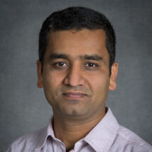 Person with short dark hair wearing a light collared shirt against a gray background.