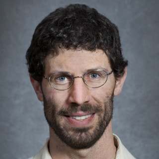 Person with short dark hair and glasses wearing a white collared shirt against a gray photo backdrop.