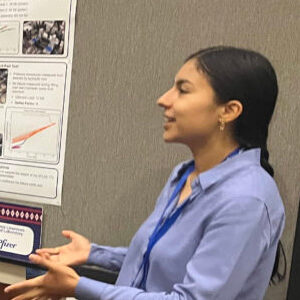 A person with long dark hair pulled back wearing a blue collared shirt talks about their research next to their poster board at a conference.