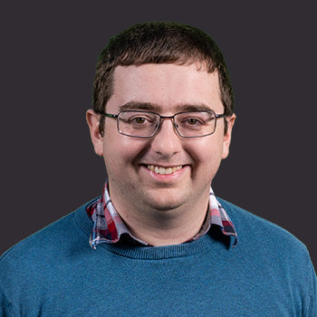 Person with short brown hair wearing glasses and a blue sweater over a red checkered collared shirt, against a black background.