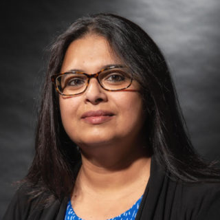 Person with medium-length black hair wearing glasses and a black cardigan over a blue top, photographed against a dark gray background.