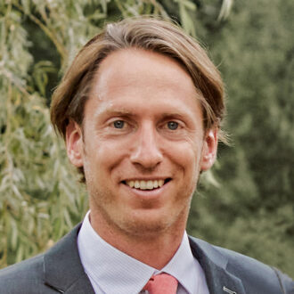 Eric Sundstrom, a person with short blonde hair wearing a gray suit over a white collared shirt and a pink tie. Photographed outdoors, with green foliage in the background.