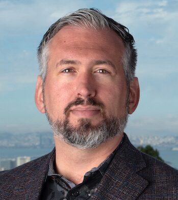 Brett Helms, a person with short gray hair and a beard wearing a dark colored blazer over a dark collared shirt, photographed outdoors, with a view of the Bay Area.