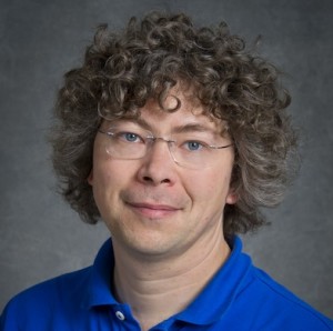Jan Kern, a person with short curly grayish hair wearing a blue collared shirt, photographed indoors against a gray backdrop.