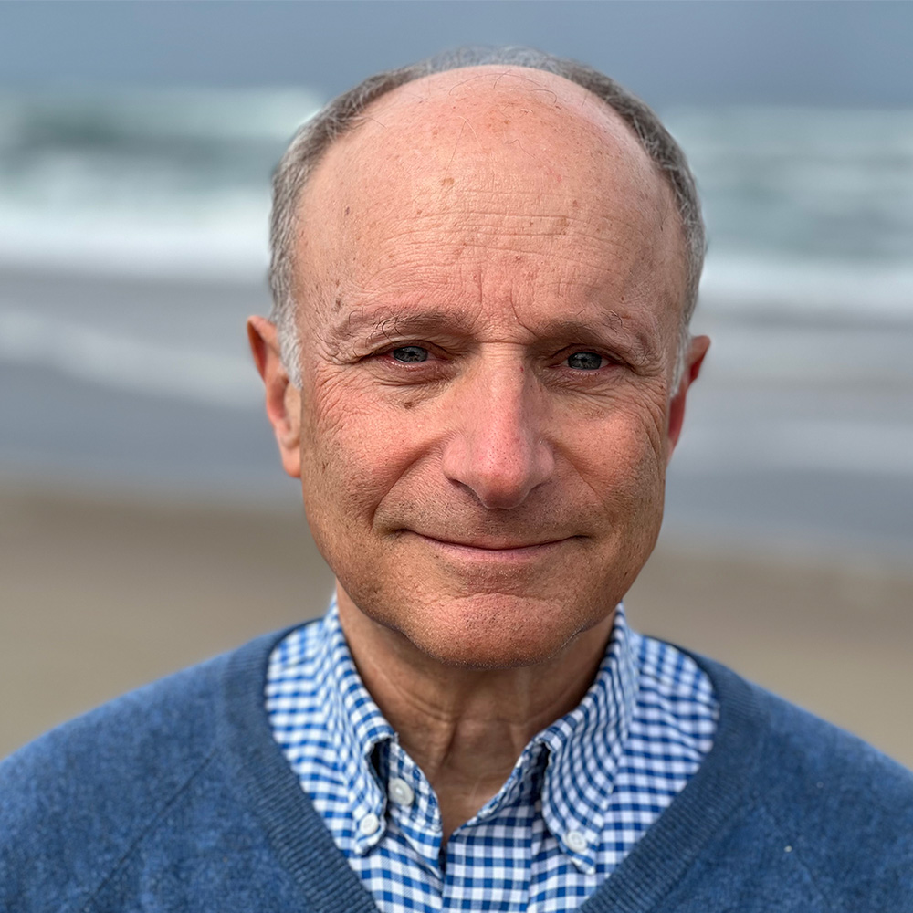 Person with gray hair wearing a navy sweater over a blue and white checkered collared shirt, photographed outdoors with an ocean in the background.