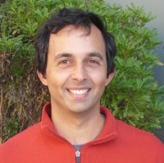 Arun Persaud, a person with short dark hair wearing a red collared shirt, photographed outdoors against a green bush.