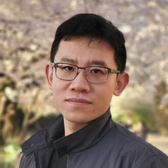 Peng Peng, a person with short dark hair, wearing glasses and a dark gray jacket over a black top, photographed outdoors with trees in the background.