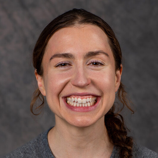 Katie Klymko, a person with long brown hair pulled back into a braided ponytail, wearing a gray shirt. Katie is photographed indoors against a gray backdrop.