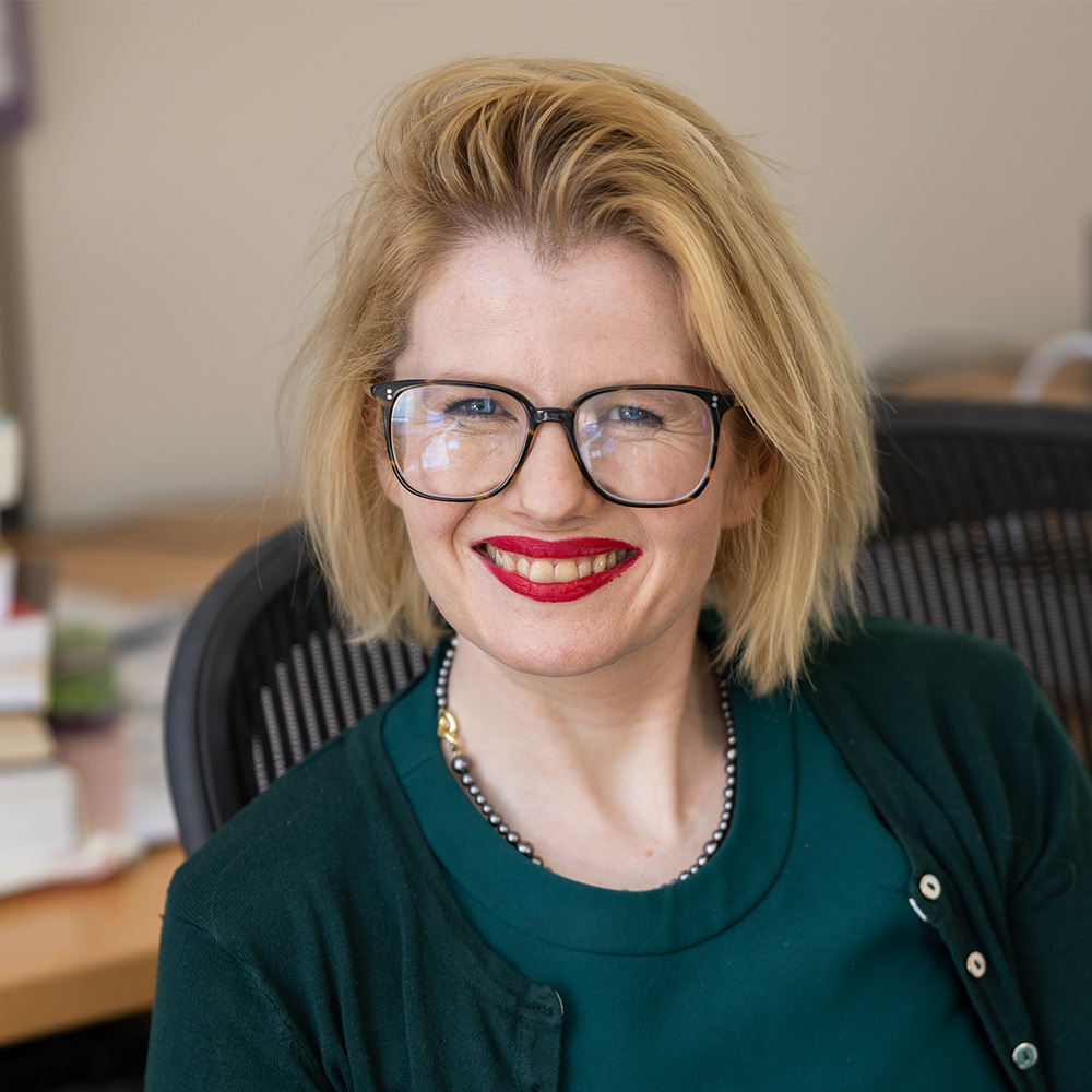 Person with short blonde hair wearing black framed glasses and a dark green cardigan over a green top. The person is seated in a chair in their office. Their desk can be seen in the background, with stacks of books, a bulletin board, and part of their computer monitor showing.