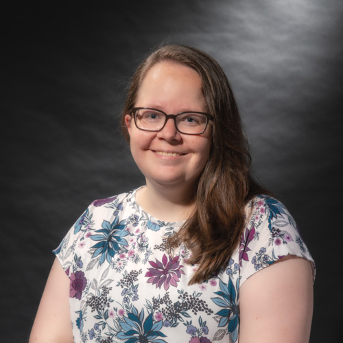 Heather Crawford, a person with long brown hair wearing a white top with a floral print. Heather is photographed against a dark gray backdrop.