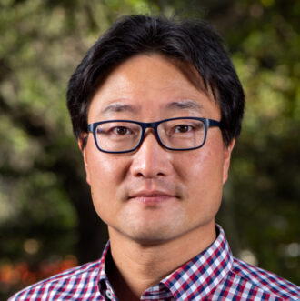 Taek Soon Lee, a person with short black hair wearing glasses and a red and white checkered collared shirt. Photographed outdoors with green foliage in the background.