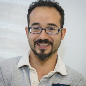 Héctor García Martín, a person with short dark hair and a beard wearing glasses and a gray and white collared top, photographed against a gray background.