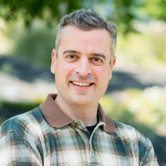 Nathan Hillson, a person with short gray hair wearing a green, brown, and white plaid shirt, photographed outdoors.