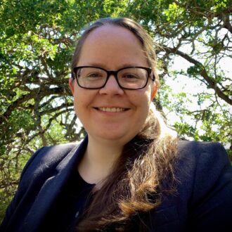 Heather Crawford, a person with glasses and long brown hair pulled back into a ponytail, wearing a dark blazer over a black top. Photographed outdoors, with trees in the background.