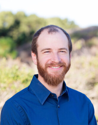 Joseph (Joe) Rand, a person with short brown hair and a beard wearing a navy collared top. Photographed outdoors against hills with green foliage.