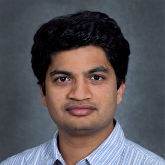 Amol Phadke, a person with short black hair wearing a white and blue striped collared top. Photographed indoors against a dark gray backdrop.