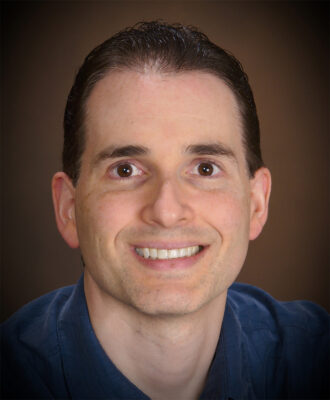 Ronnen Levinson, a person with short dark hair wearing a navy shirt, photographed against a dark copper background.