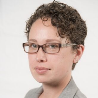 C. Anna Spurlock, a person with short curly brown hair wearing glasses and a gray blazer over a black top, photographed against a light gray background.